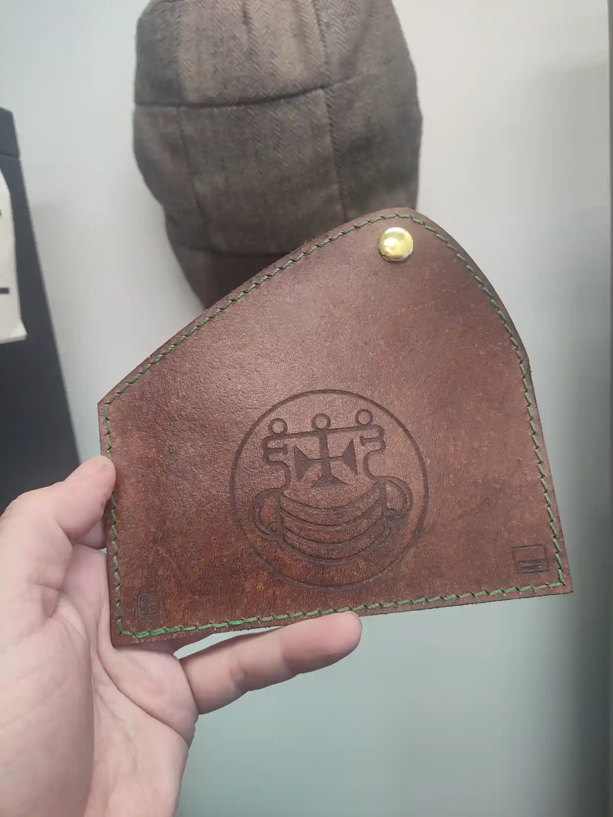 A hand holds a brown leather wallet with green stitching and a brass rivet, featuring an embossed circular logo that…