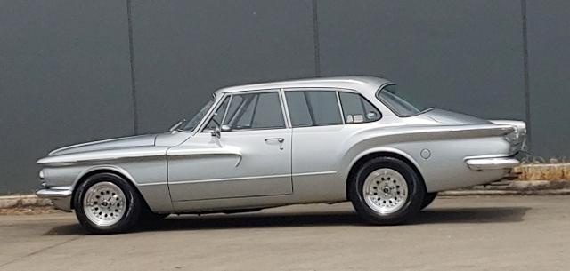 a silver vintage four-door sedan with a distinctive curved roofline and chrome trim is parked against a plain gray wall. The…