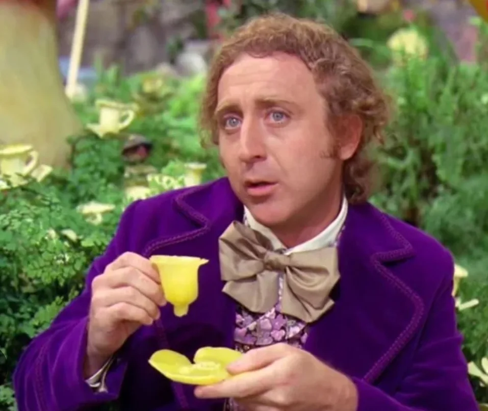 a man with curly hair and a purple velvet jacket holds a yellow teacup and saucer, looking shocked with his mouth open. The…