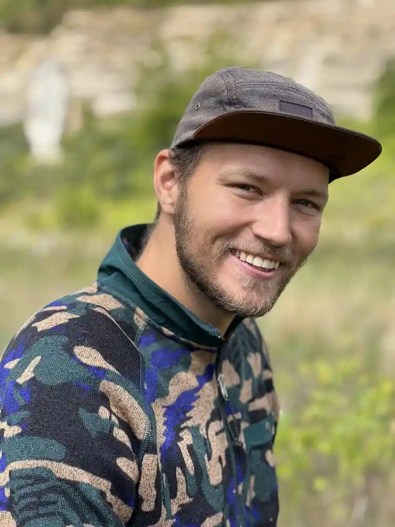 A man with a beard and a grey baseball cap smiles at the camera while wearing a zip-up jacket with a blue, green, and beige…
