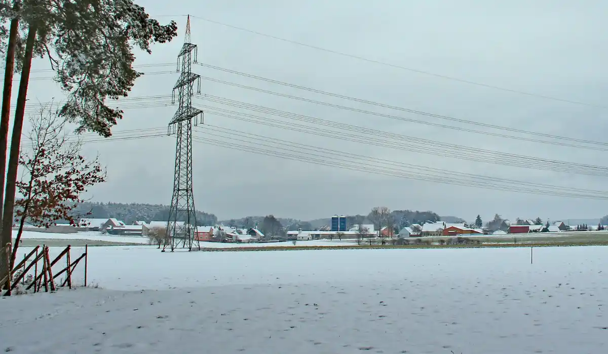 A snowy rural landscape with a tall electricity pylon in the middle ground, power lines stretching across the sky, and…
