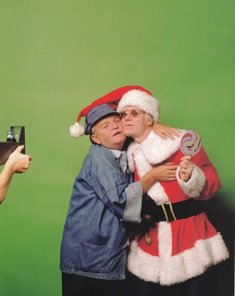 Two men pose against a green screen; one wears a denim jacket and blue cap with a red Santa hat perched on his head, while…