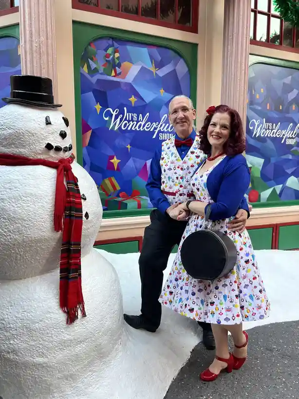 A man and woman in matching festive holiday-themed outfits pose for a photo next to a large snowman statue wearing a top hat…