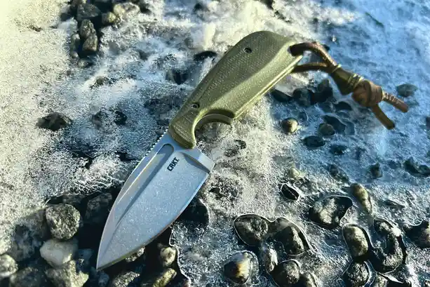 A CRKT brand pocket knife with a green textured handle and leather lanyard rests on a bed of wet, frosted rocks. The blade…
