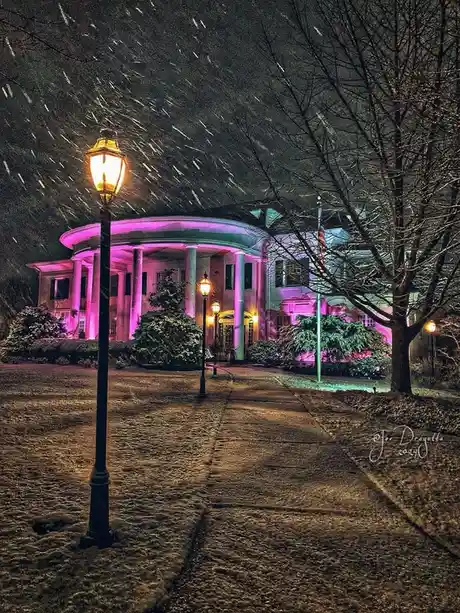 A nighttime photograph of the White House illuminated with vibrant pink and purple lights during a heavy snowfall, with…