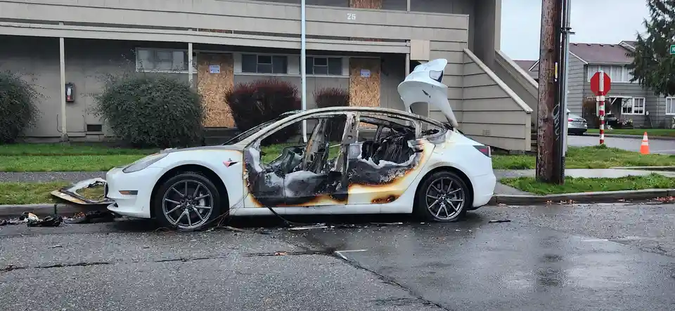 A white Tesla Model 3 is severely burned and destroyed, with its interior charred, doors and hood open, and the front bumper…