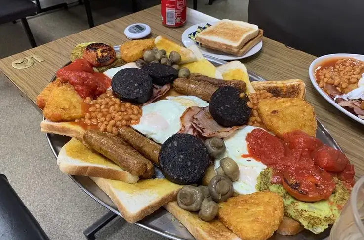 a large, overflowing plate of a traditional full english breakfast featuring fried eggs, bacon, sausages, black pudding…