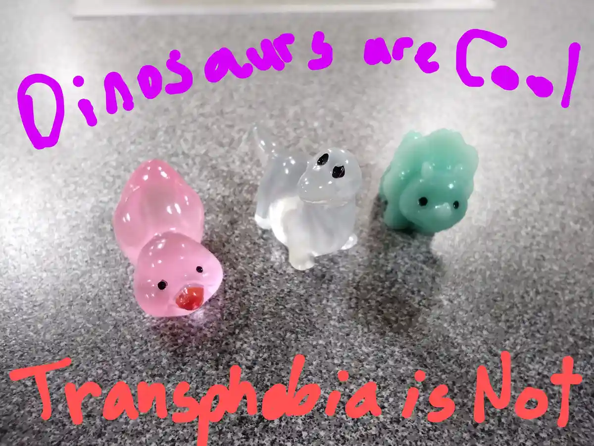 Three small, translucent plastic dinosaur figurines — pink, white, and green — sit on a speckled gray surface. Purple text…