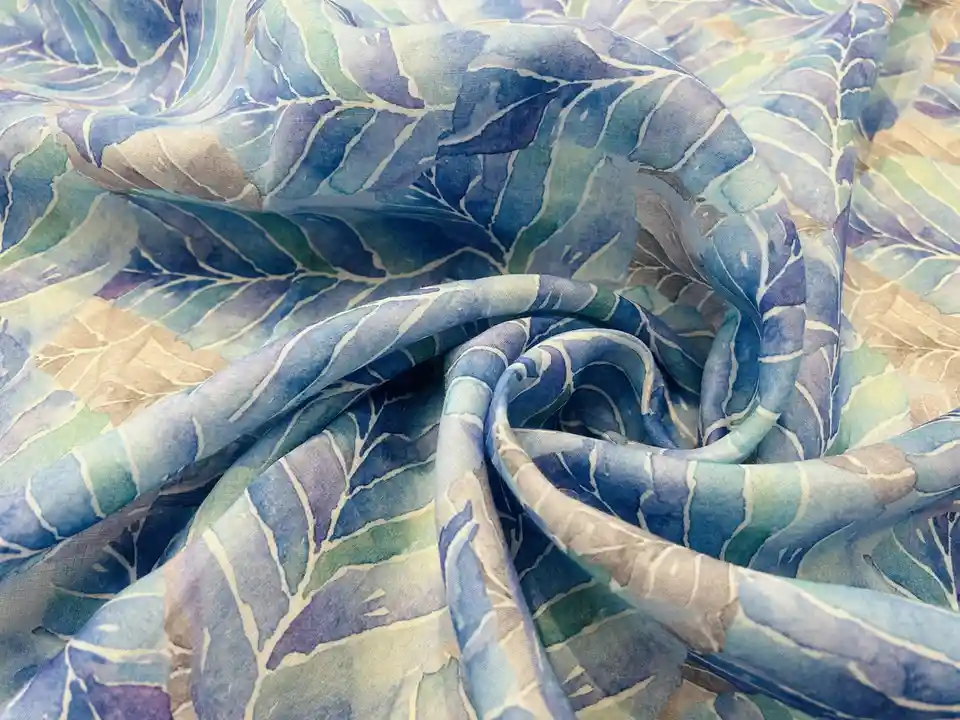 A close-up photo of a soft, flowing scarf with a watercolor-style leaf pattern in shades of blue, green, and beige. The…