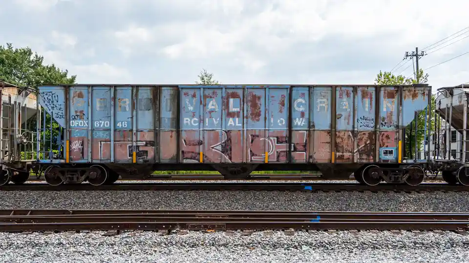 A rusty, weathered blue grain train car sits on tracks under a cloudy sky, with faded lettering spelling 'PERIVAL GRAIN'…