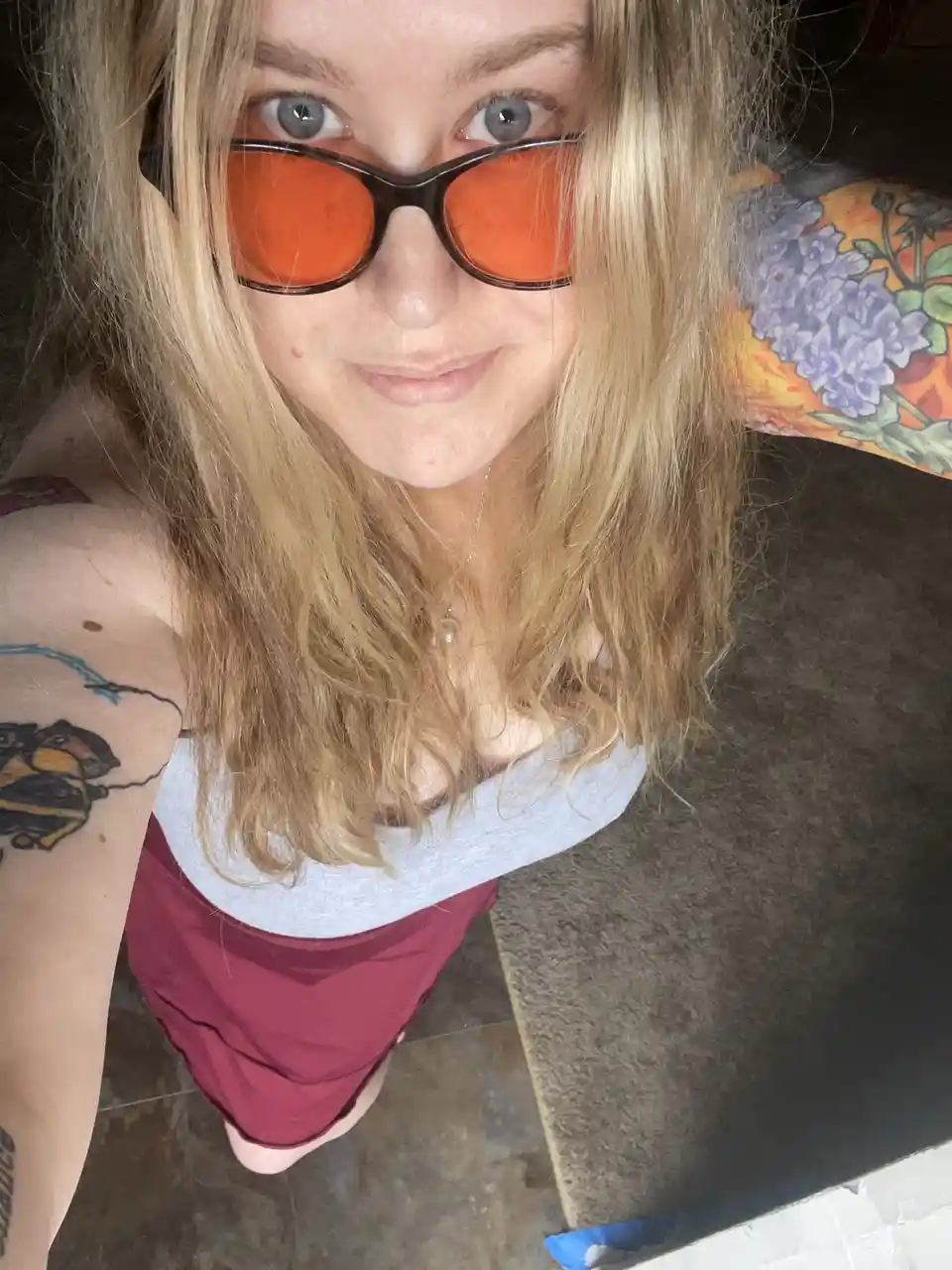 A selfie of a person with blonde hair and blue eyes wearing bright orange-tinted sunglasses, looking directly at the camera…