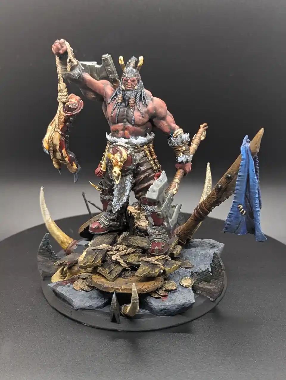 A highly detailed, painted miniature of a muscular, horned demon-like figure with a long beard, holding a bloody, hanging…