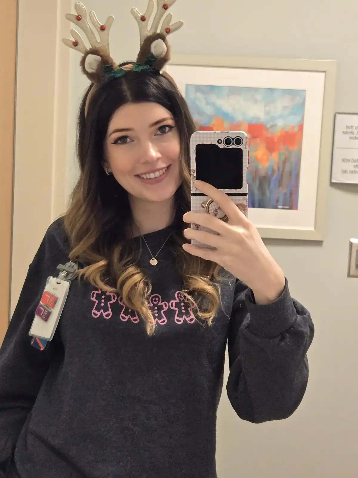 A woman in a dark grey sweatshirt with pink gingerbread man outlines smiles while taking a mirror selfie, wearing reindeer…