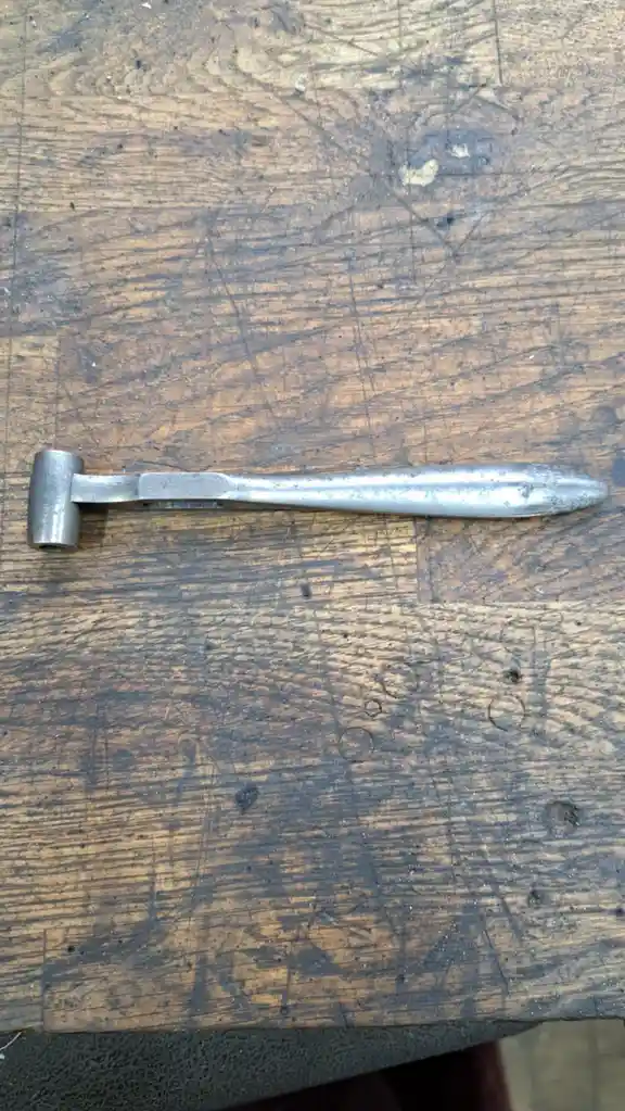 A metal hammer with a rounded head and a tapered, pointed handle lies on a heavily scratched wooden workbench. The overlaid…