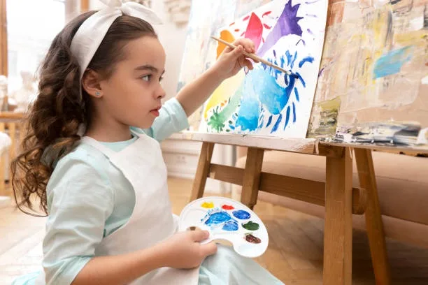 A young girl with curly hair and a white headband is painting on a canvas with a brush, holding a paint palette in her other…
