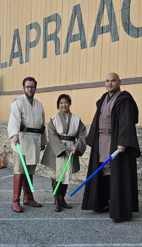 Three people dressed as Star Wars Jedi, holding lightsabers, stand in front of a wooden wall with the word 'LAPRAA' painted…