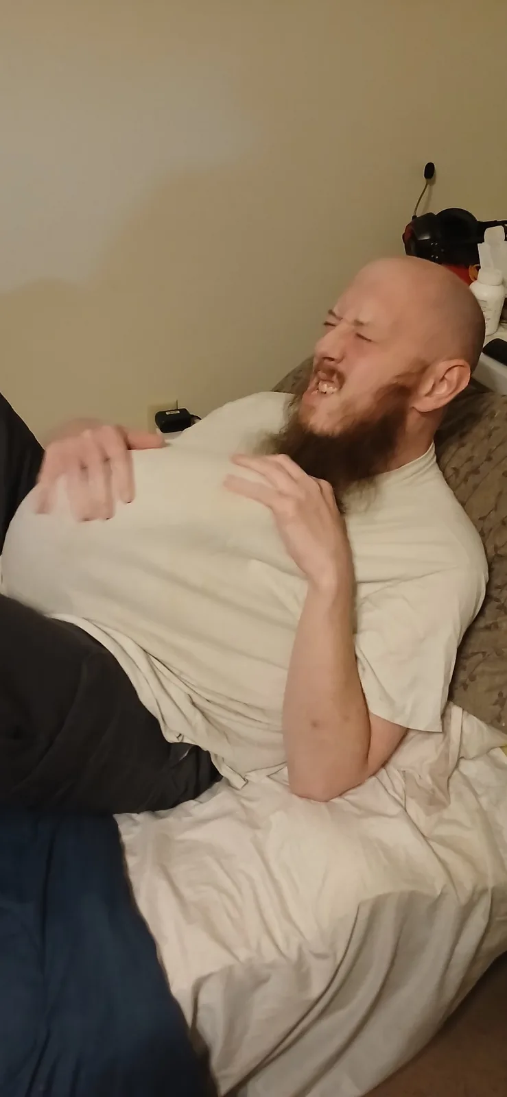 A bald man with a long beard is lying on a bed, clutching his stomach and grimacing in apparent agony or intense laughter…