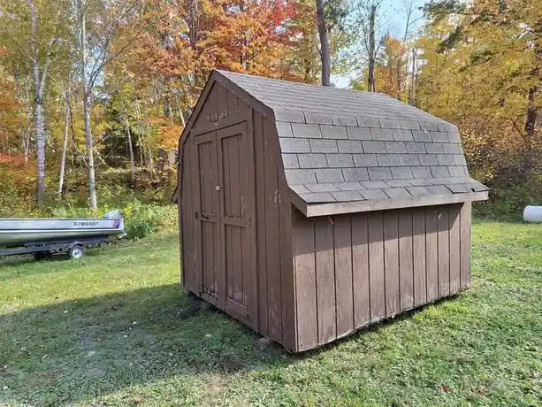 A brown wooden shed with a shingled roof sits on a grassy lawn surrounded by autumn trees. To the left, a boat on a trailer…