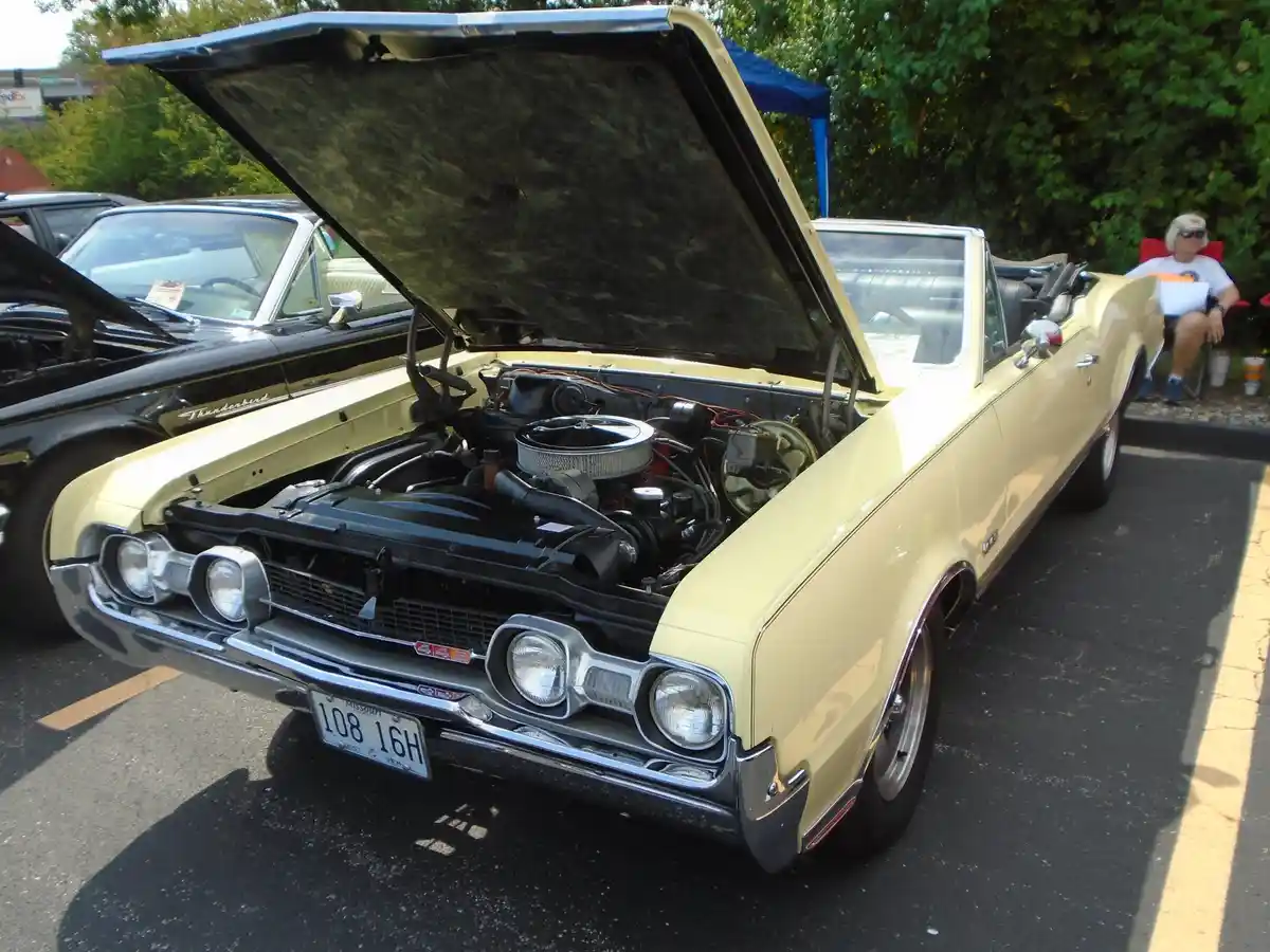 A pale yellow classic convertible with its hood open, revealing a detailed engine bay, parked at a car show. A person sits…