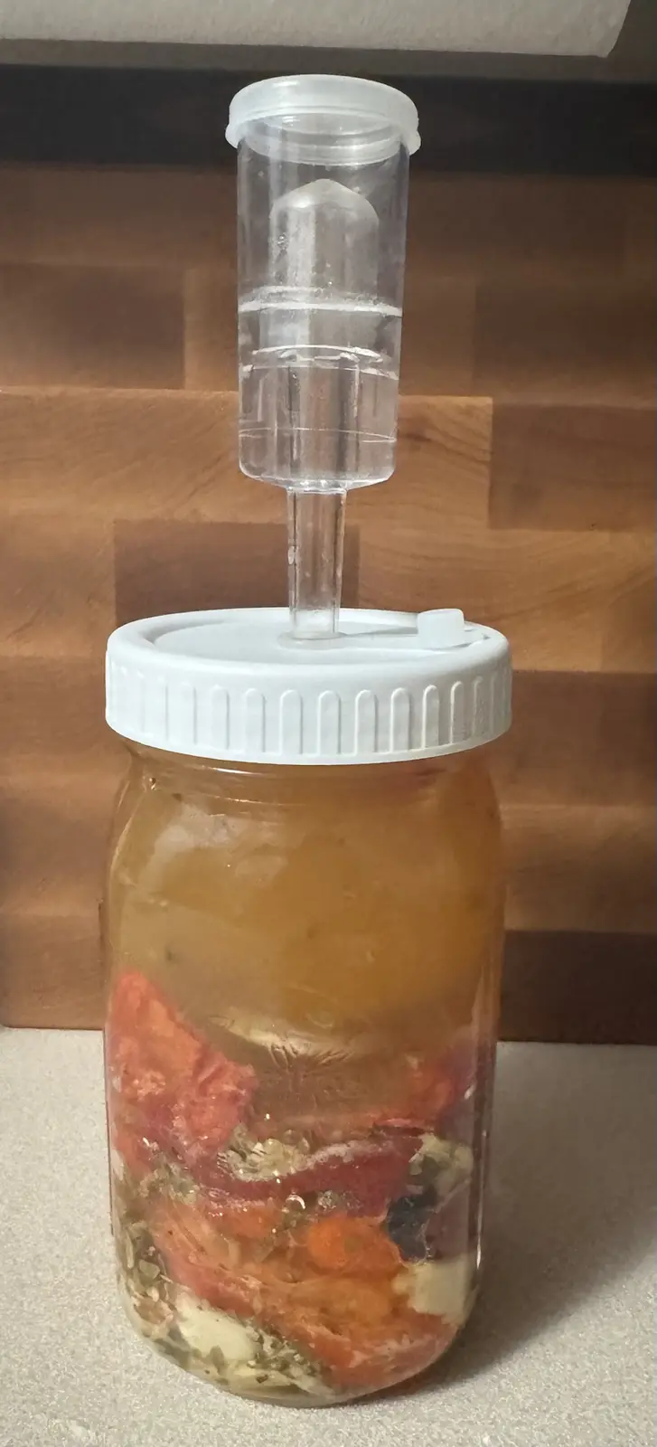 A glass mason jar filled with sliced red vegetables and brine, topped with a white lid and a clear plastic airlock tube…