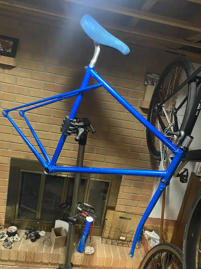 A bright blue bicycle frame is mounted on a repair stand in a garage with a brick wall and fireplace. The frame has a blue…