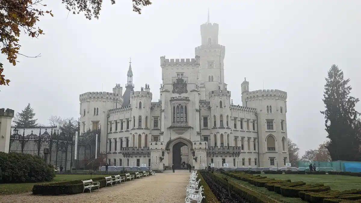 A large, ornate, light-colored castle with multiple towers and turrets stands under an overcast sky, flanked by manicured…