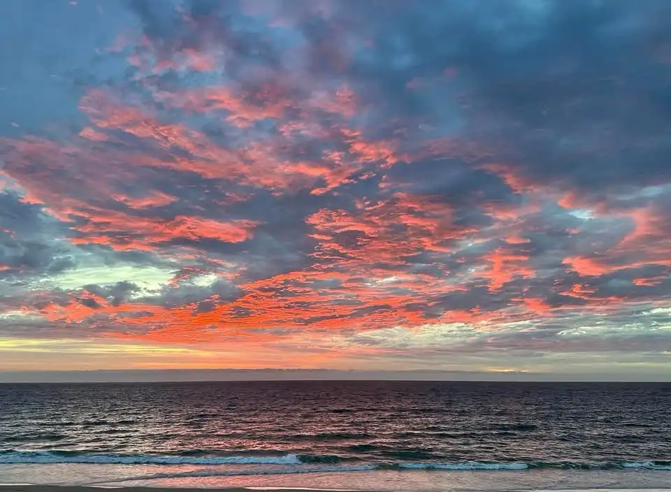 A serene beach scene at sunset or sunrise, with vibrant pink, orange, and blue clouds reflecting over dark ocean waters. The…