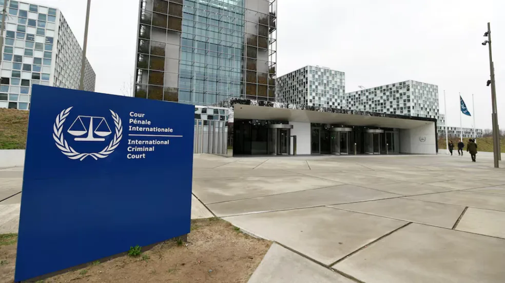 A blue sign with the International Criminal Court logo and text stands in front of a modern glass building complex. The sign…