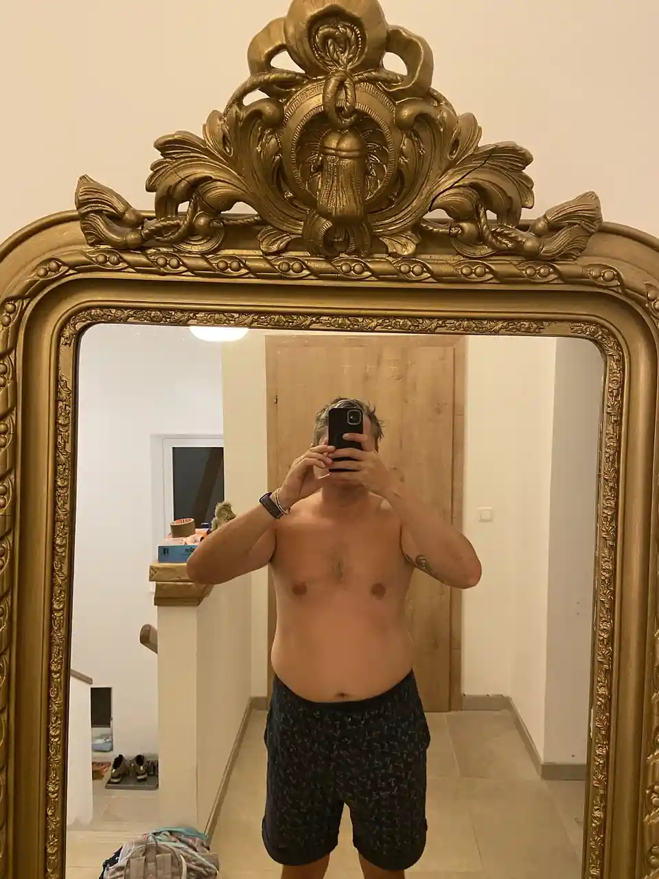 A shirtless man with a visible belly stands in front of an ornate, gilded mirror, holding a phone to obscure his face. The…