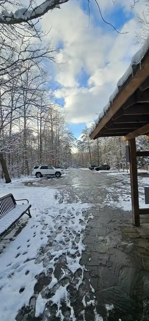 A snowy outdoor scene with a stone path partially covered in snow and ice, leading toward a parking area with cars. Bare…