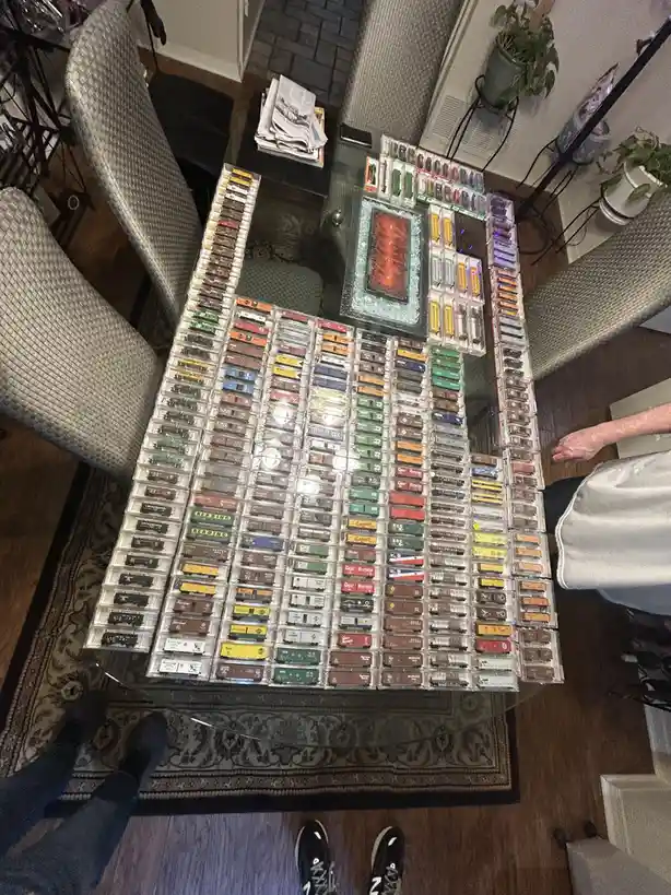 An overhead shot shows a glass-top table covered with hundreds of model train cars in clear plastic cases, arranged in neat…