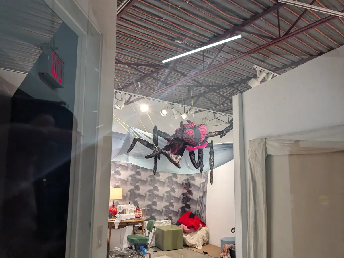 A large, decorative spider sculpture with a pink and black patterned body and black lace-like legs hangs from the ceiling of…