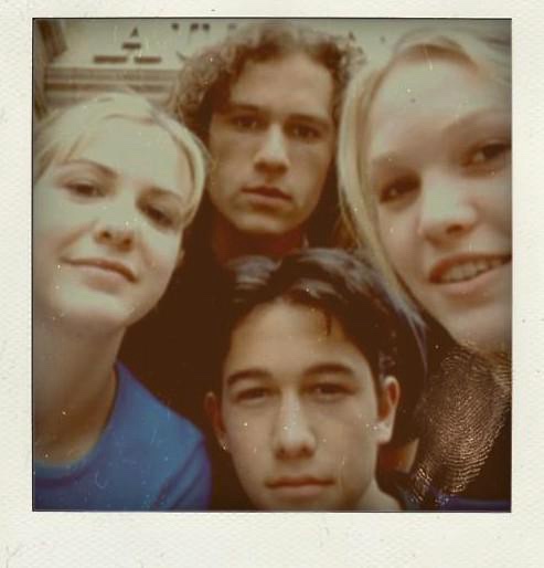 A grainy, vintage-style photo shows four young people posing closely together, with one person in the center looking…