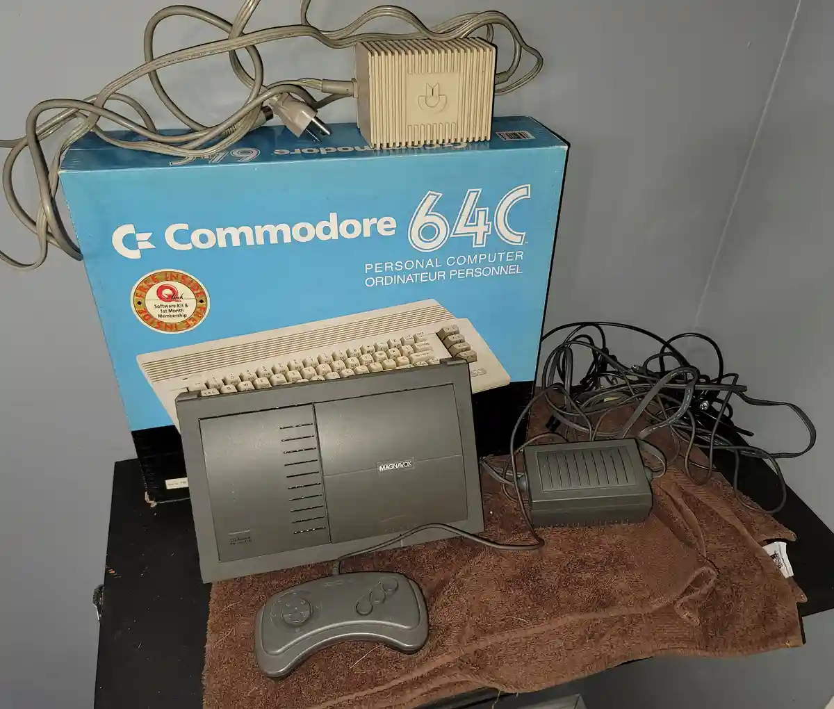 A vintage Commodore 64C personal computer box sits behind a Magnavox monitor, a grey game controller, and a tangle of cables…