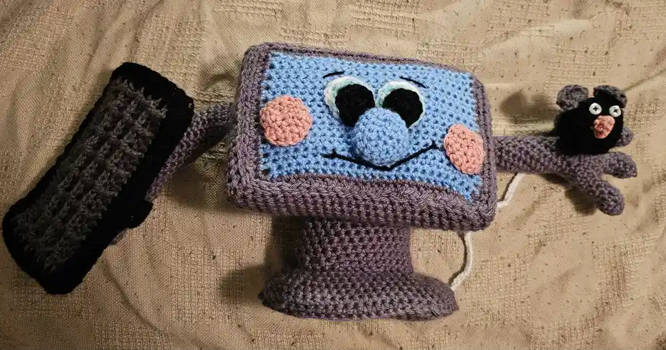 A handmade crocheted computer monitor with a smiling blue face, pink cheeks, and black eyes sits on a beige fabric surface…