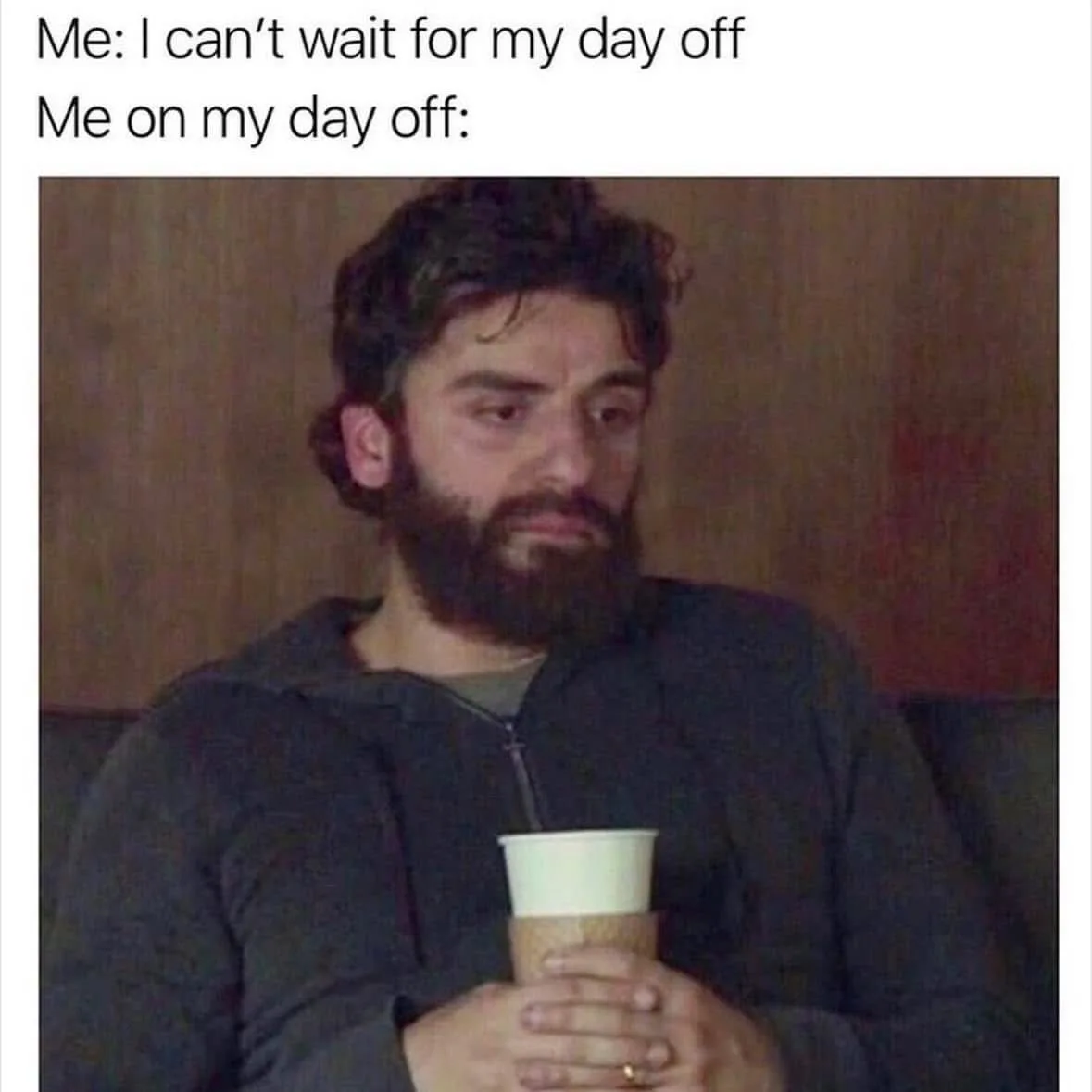A man with a beard and dark hair, wearing a dark hoodie, sits holding a coffee cup with both hands. He has a weary…