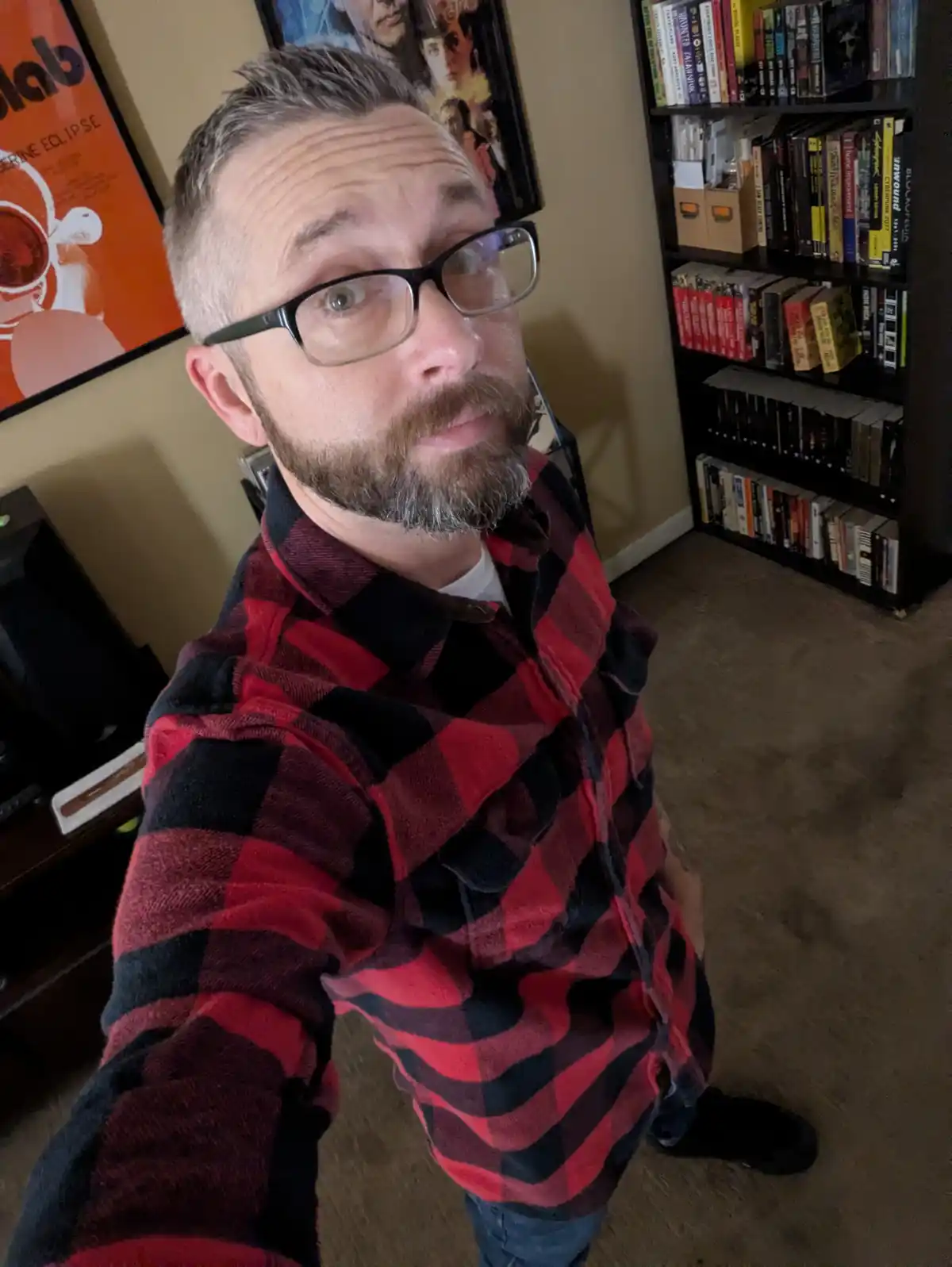 A man with glasses and a beard wearing a red and black plaid shirt takes a selfie in a room with bookshelves and posters on…