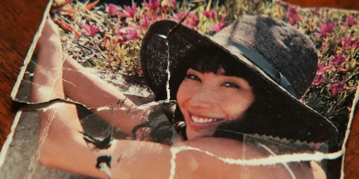 A visibly torn and damaged old photograph shows a smiling woman with dark hair and a wide-brimmed hat, lying in a field of…