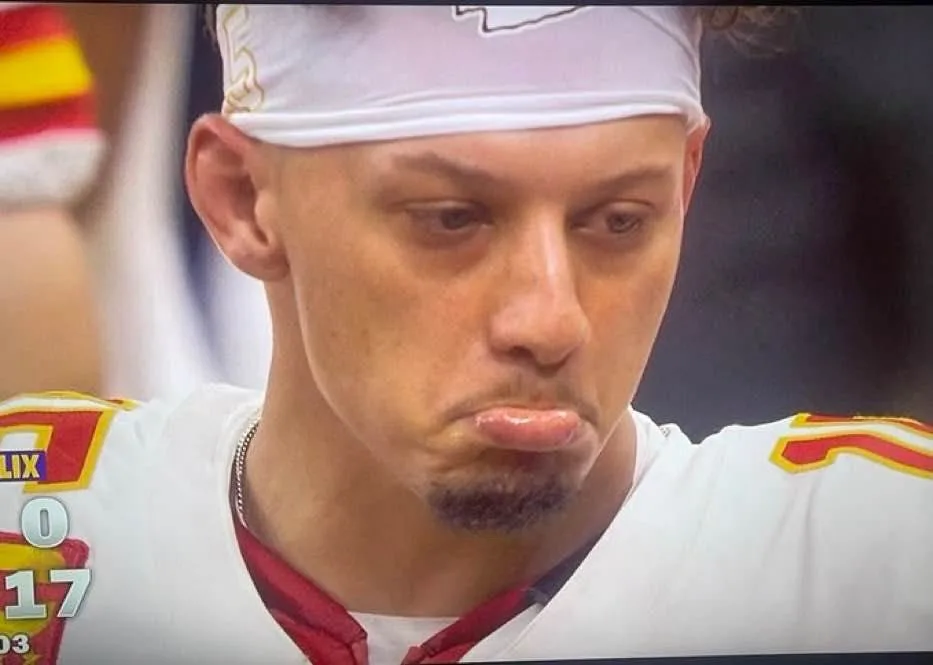 A close-up of a football player wearing a white jersey with red and yellow trim and a white headband, looking down with a…