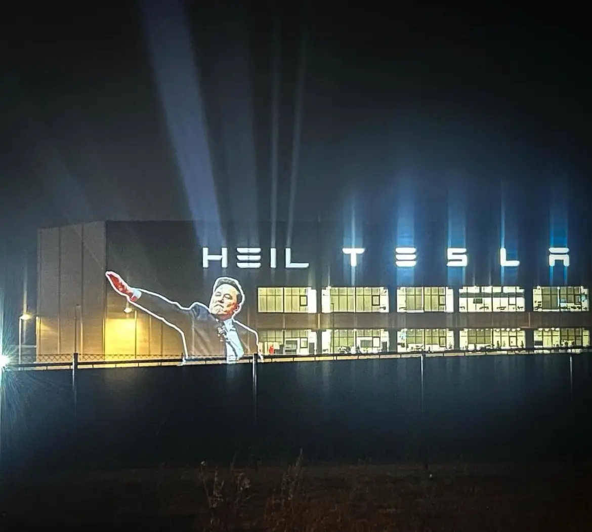 A nighttime photo of a large industrial building with illuminated windows and spotlights. Projected onto the side of the…