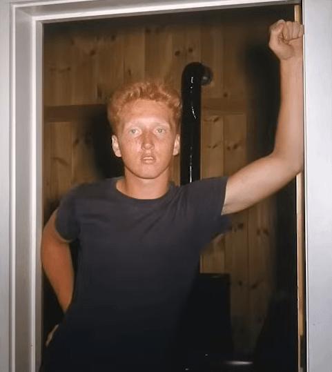 A man with red hair and a black t-shirt stands in a doorway, raising his right fist in a triumphant or defiant gesture…