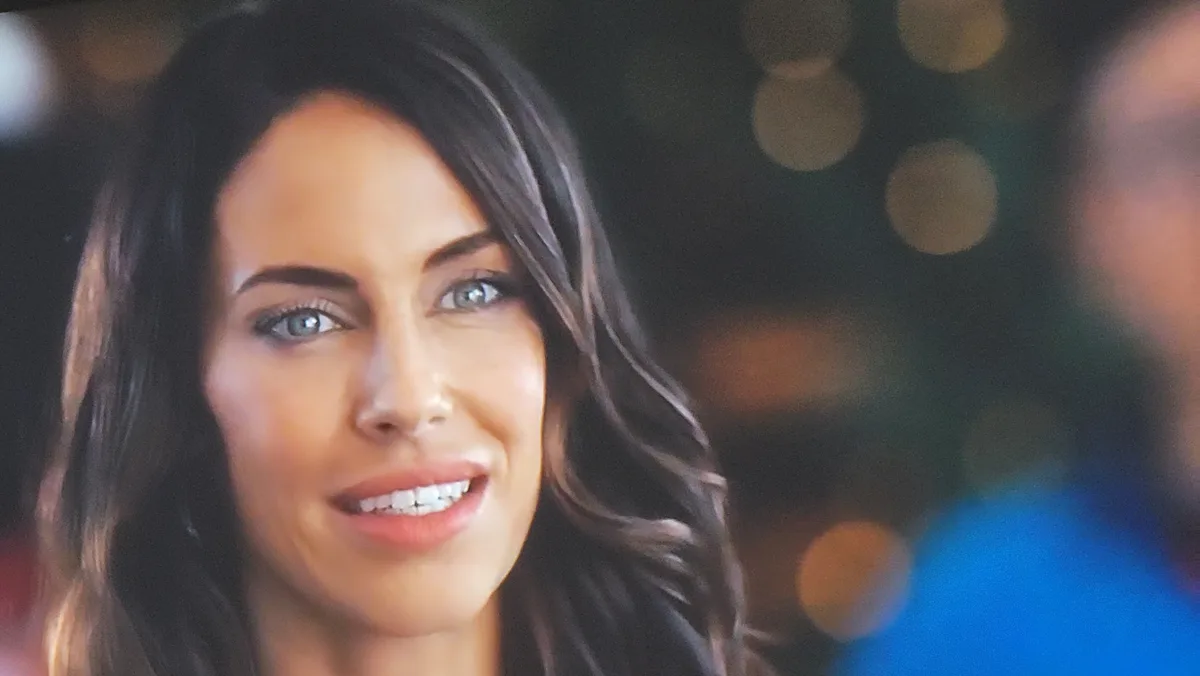A close-up image of a woman with dark hair and blue eyes smiling slightly while looking forward, with a blurred background…