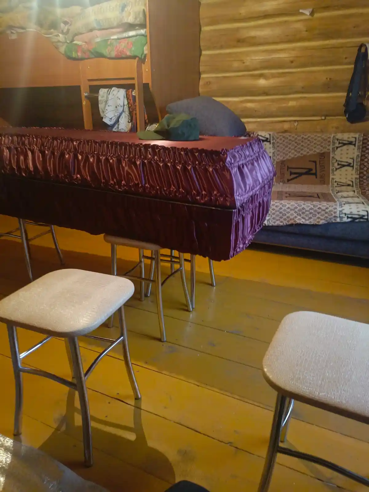 A coffin draped in a shiny purple and burgundy fabric cover sits on metal stools in a rustic room with wooden walls and…