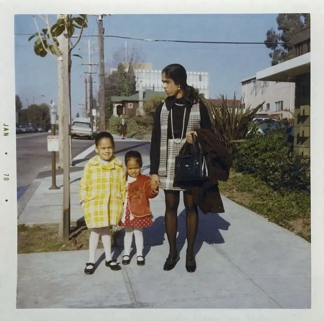 A vintage 1970s photo shows a woman holding hands with two young girls on a sidewalk. The older girl, wearing a yellow plaid…
