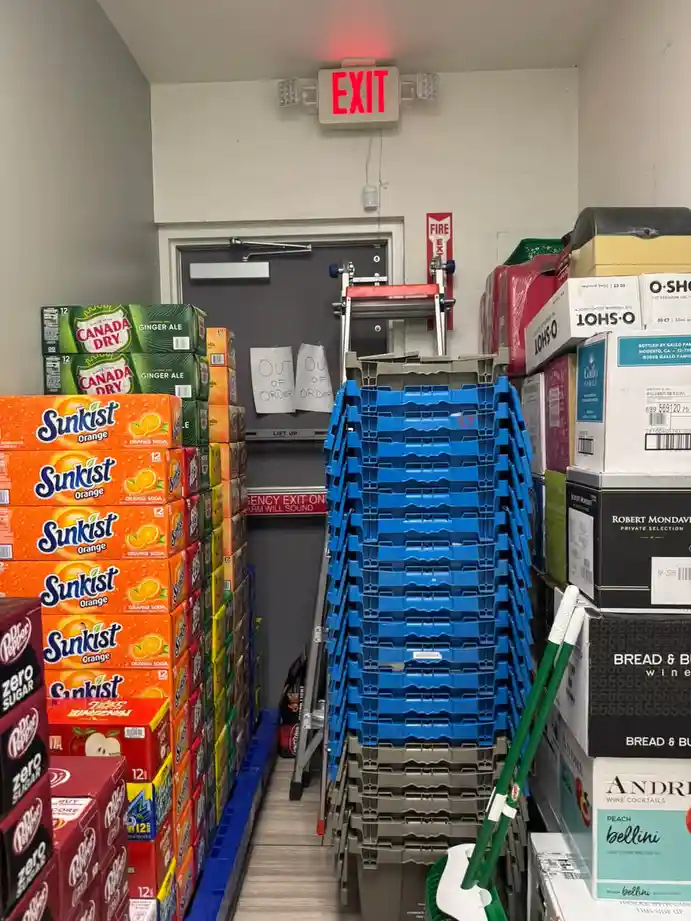A narrow store aisle is blocked by stacks of soda cases, blue plastic crates, and a ladder, with a red 'EXIT' sign above a…