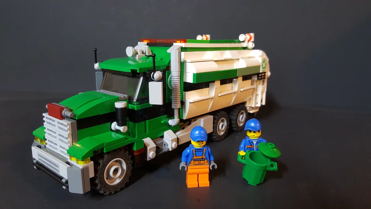 A green and beige LEGO garbage truck with two LEGO minifigures in blue uniforms and hard hats standing beside it. One…