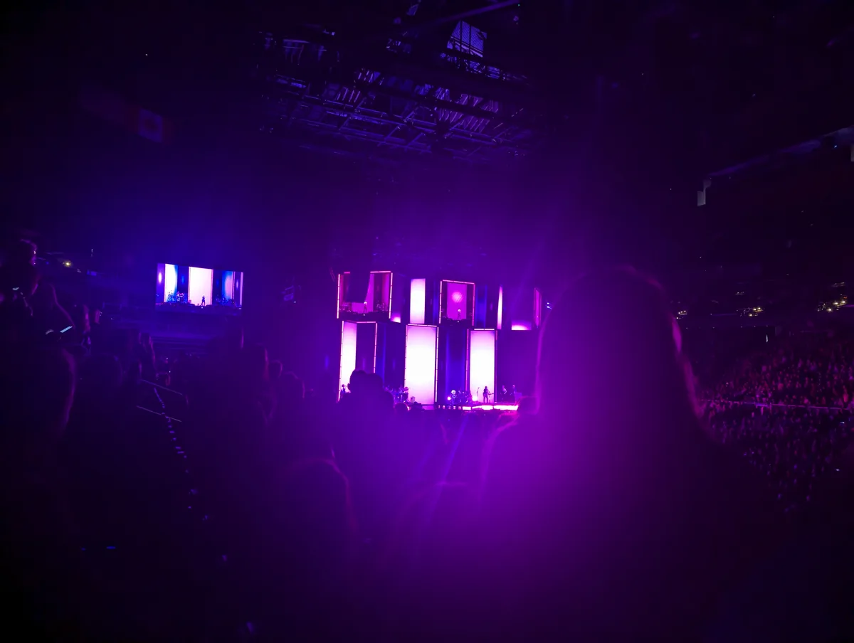 A dimly lit concert venue bathed in purple light, showing a stage with performers and large screens, viewed from the…