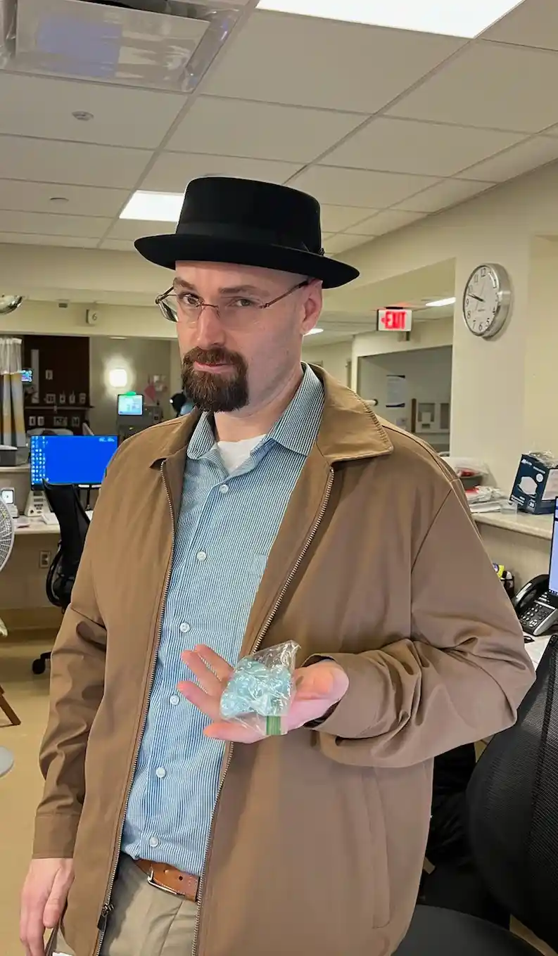 A man in an office wearing a black fedora, glasses, and a brown jacket holds a small, crumpled blue plastic item in his…