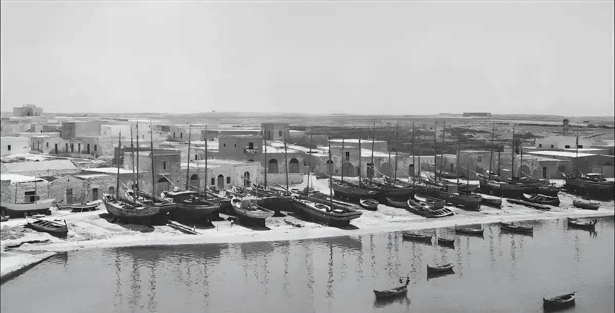 A black-and-white historical photograph shows a quiet harbor with numerous wooden boats resting on a sandy shore, some…