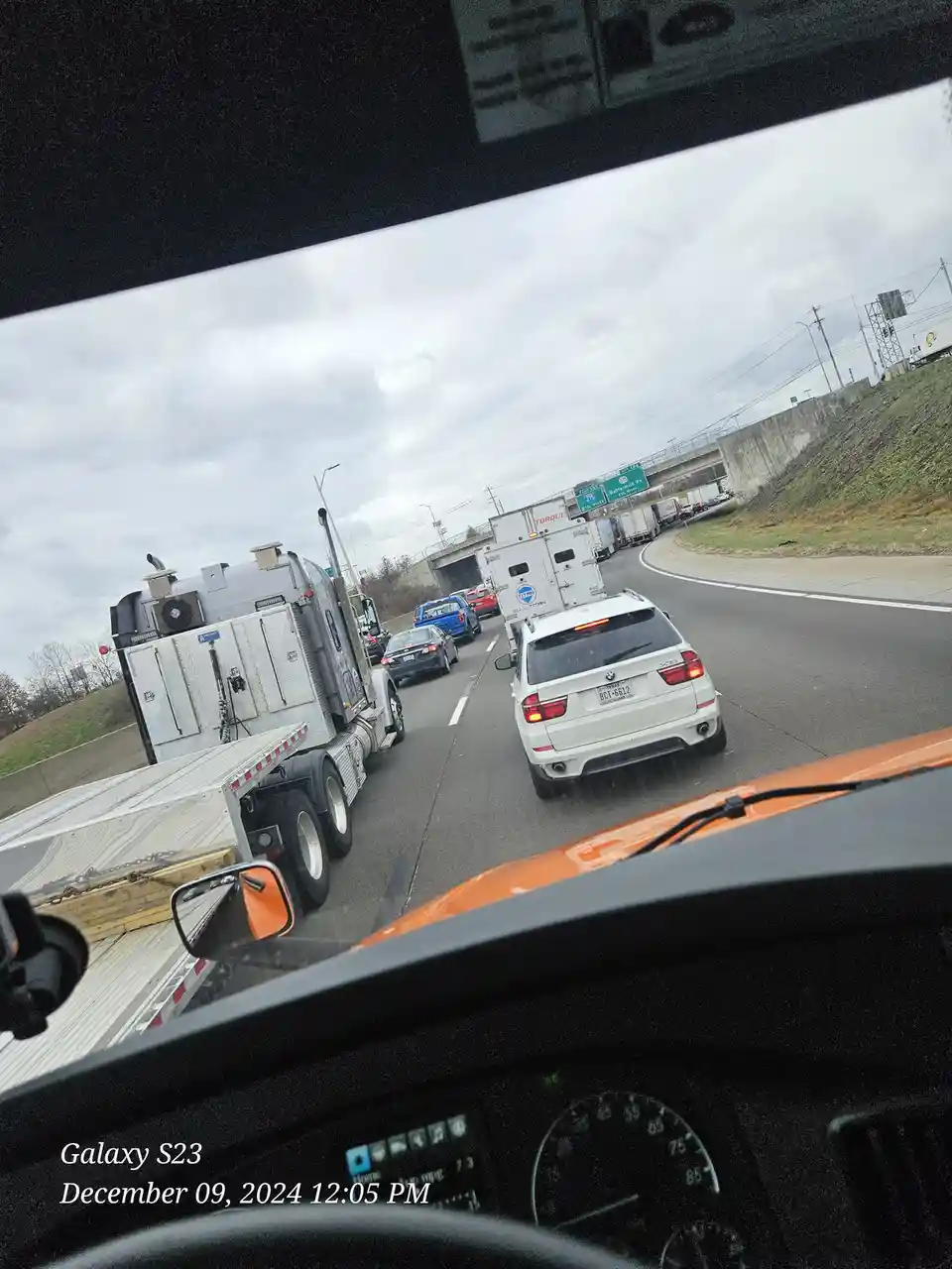 A dashcam photo from inside an orange car shows heavy traffic on a highway with a large silver semi-truck and a white BMW X5…